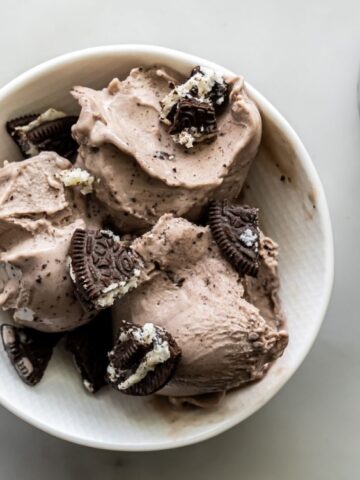 A white bowl filled with scoops of chocolate ice cream topped with crumbled chocolate sandwich cookies sits on a light surface next to a container of more ice cream.