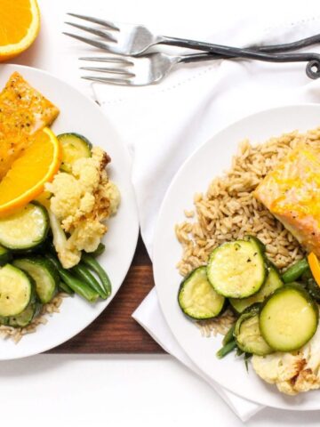Two plates with salmon fillets, brown rice, zucchini, cauliflower, snap peas, and orange slices are on a wooden board, with forks, napkin, and orange halves beside them.