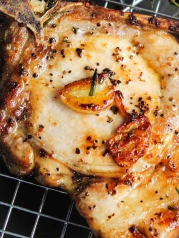 A cooked pork chop with garlic, herbs, and cracked black pepper sits on a metal cooling rack.