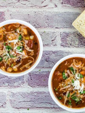 Two bowls of soup topped with shredded cheese and herbs, placed on a brick background, with two slices of bread and two metal spoons beside them.