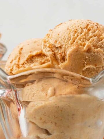 Two scoops of light brown ice cream served in a clear glass bowl, with another bowl of ice cream blurred in the background.