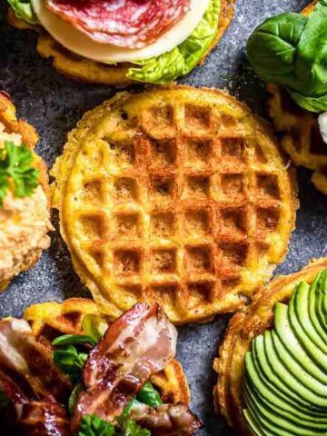 Five savory waffles are topped with various ingredients, including avocado, bacon, spinach, tomato, cheese, lettuce, and a fried egg, arranged around a plain waffle on a dark surface.