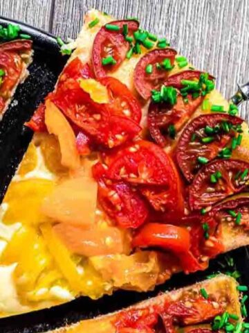 A slice of tomato and cheese tart topped with chives is being lifted from a black pan on a wooden surface.