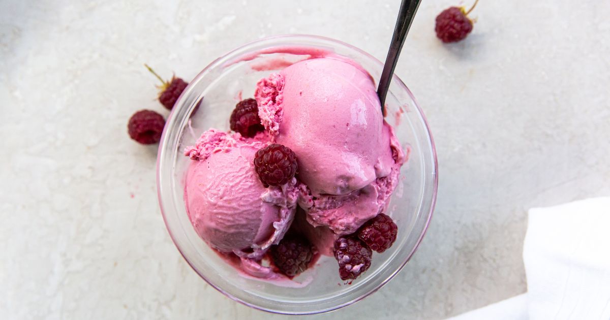 A glass bowl with three scoops of pink raspberry ice cream, garnished with whole raspberries, sits on a light surface with a spoon in the bowl.