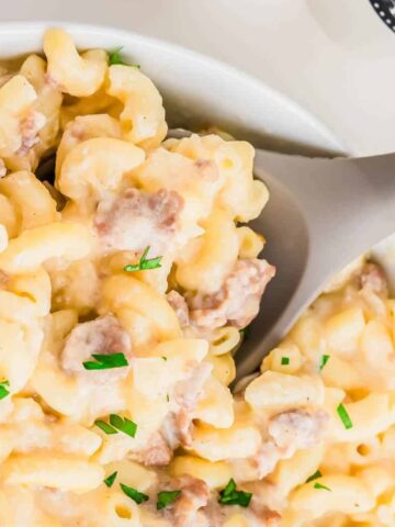 A bowl of macaroni and cheese with ground meat, garnished with chopped herbs, being served with a gray and wooden spoon.