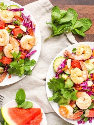 Two plates of shrimp salad with cucumbers, red cabbage, watermelon, and herbs sit on a wooden table; a bowl of watermelon slices and mint leaves are beside them.