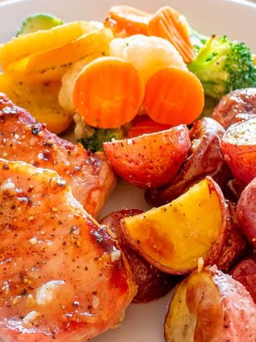 A plate of seasoned pork chops, roasted red potatoes, and steamed mixed vegetables including carrots, broccoli, zucchini, and yellow squash.