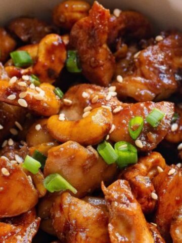 A bowl of stir-fried chicken with cashews, garnished with chopped green onions and sesame seeds.