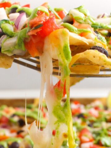 A close-up of a fork lifting cheesy nachos topped with black beans, diced tomatoes, onions, and jalapeños, with melted cheese stretching from the chips.
