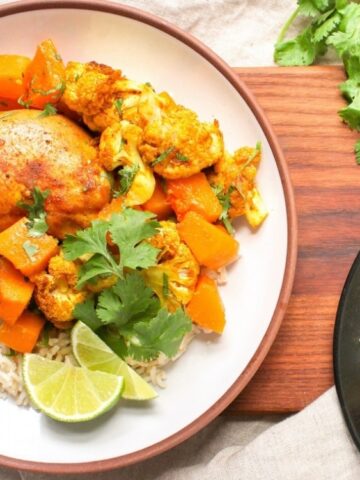 A plate of rice topped with roasted chicken, cauliflower, and orange vegetables, garnished with cilantro and lime wedges. Another plate with a similar dish is partially visible.