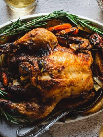 A whole roasted chicken with crispy skin is served on a platter with roasted vegetables and garnished with fresh rosemary.