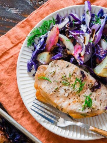 A plate with a grilled pork chop, roasted potatoes, and sautéed red cabbage and apples, garnished with dill, on an orange napkin with a fork.