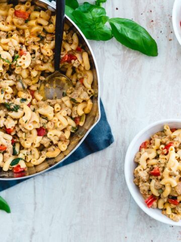 A pot and two bowls of macaroni mixed with ground meat, red bell peppers, spinach, and herbs are on a white surface with fresh basil leaves nearby—perfect inspiration for easy skillet recipes.