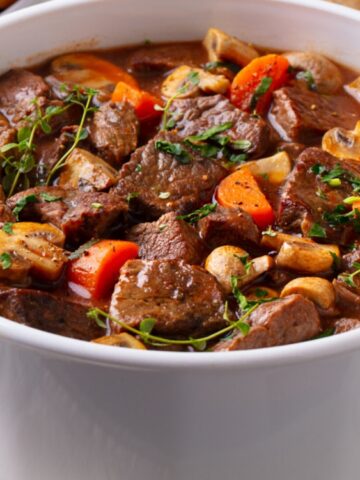 Beef stew with chunks of beef, carrots, mushrooms, and herbs in a white casserole dish.