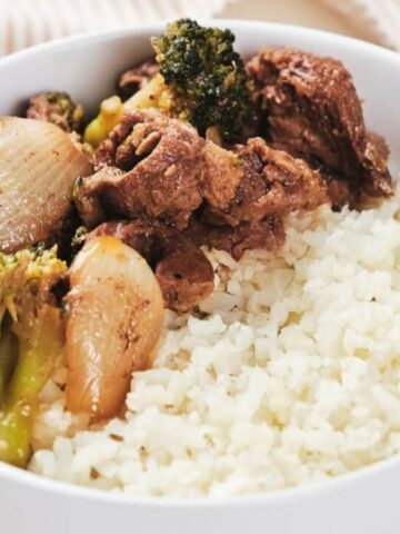 A white bowl contains cauliflower rice topped with cooked beef, broccoli, and onions.