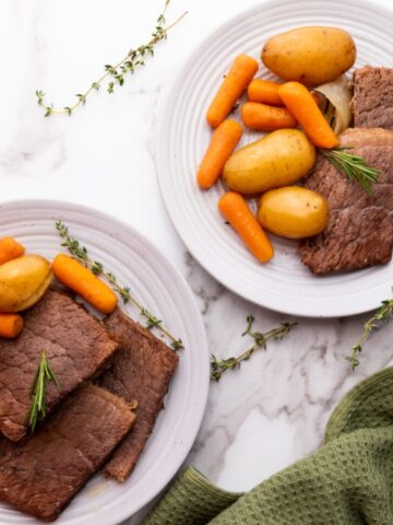 Two white plates each with slices of roast beef, baby potatoes, and carrots, garnished with rosemary, on a marble surface with a green cloth and herbs.