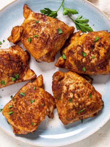 A plate with five seasoned, roasted chicken thighs garnished with chopped parsley, next to a beige napkin and parsley sprigs.