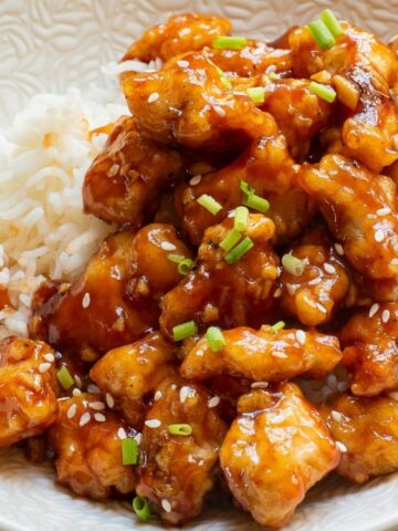 A bowl of white rice topped with glazed chicken pieces, garnished with sesame seeds and chopped green onions.