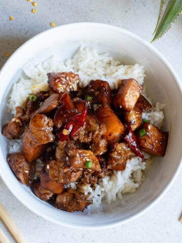A bowl of white rice topped with stir-fried chicken and red chili peppers, with chopsticks and a bamboo mat nearby on a light surface.
