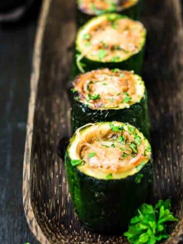 Four pieces of stuffed zucchini are arranged in a wooden tray, garnished with chopped herbs, on a dark surface.