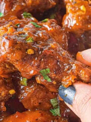 Close-up of a hand picking up a piece of saucy, garnished meat or chicken from a pile, with visible herbs and spices on the food.
