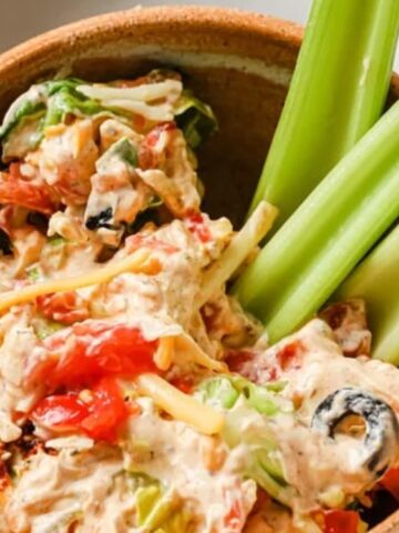 A bowl of creamy dip with chopped vegetables and olives, served with three celery sticks.