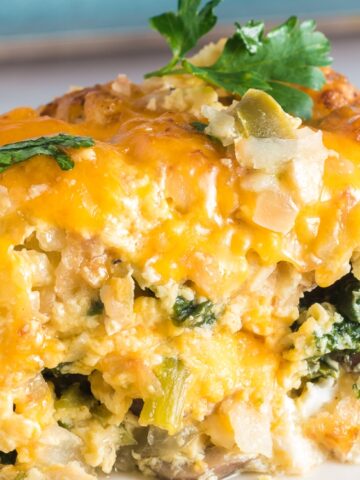 A slice of baked casserole with melted cheese, leafy greens, mushrooms, and herbs, served on a white plate.
