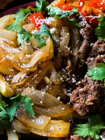 A plate with grilled onions, sliced beef, avocado, red bell peppers, fresh cilantro, and crumbled cheese.