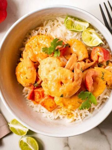 A bowl of shrimp curry with white rice, garnished with lime wedges and cilantro, sits on a marble surface next to a knife and fork.