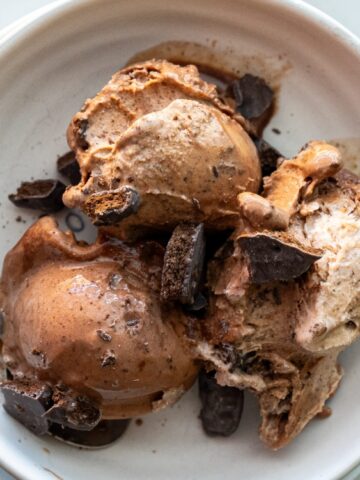 Three scoops of chocolate ice cream with chocolate chunks in a white bowl, with a spoon on the side.