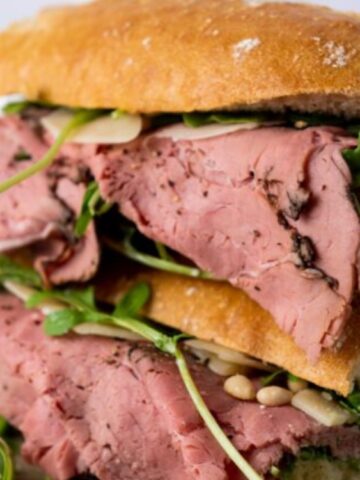 A stacked roast beef sandwich with greens and pine nuts on rustic bread, placed on a wooden surface.
