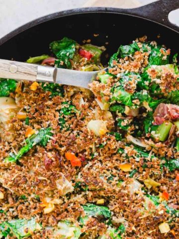 A cast iron skillet filled with sautéed greens and vegetables topped with crispy breadcrumbs, with a serving spoon resting inside.