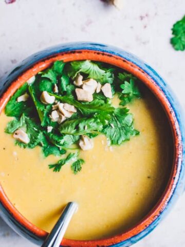 A bowl of creamy yellow soup garnished with fresh cilantro and chopped cashews, with a spoon inside and herbs scattered around on a light surface.