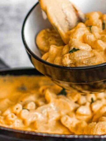 A hand serves creamy pasta from a skillet into a black-rimmed bowl, with sauce coating the pasta.