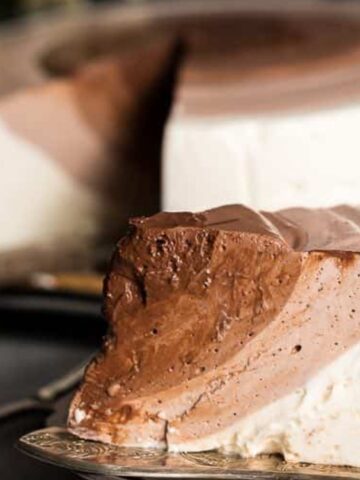 A close-up of a slice of layered chocolate and vanilla mousse cake on a plate, with the whole cake and a serving knife in the background.
