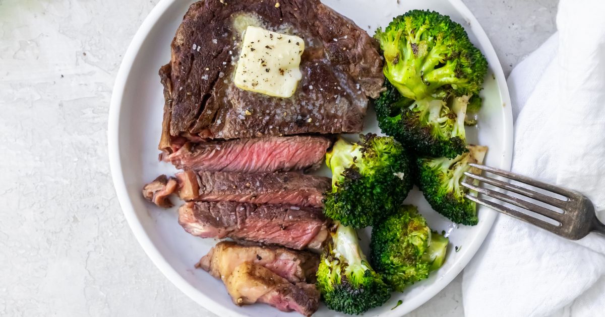 A plate with a cooked steak topped with a pat of butter, sliced, served with roasted broccoli and a fork on the side.