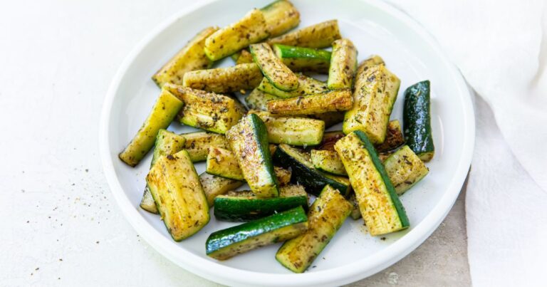 A white plate with roasted zucchini sticks seasoned with herbs and spices, placed on a light-colored surface.