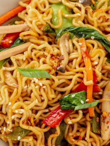 A bowl of vegetable stir-fried noodles with mushrooms, bell peppers, bok choy, and sesame seeds, with chopsticks resting on the side.
