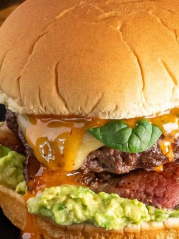 A cheeseburger with spinach, bacon, melted cheddar, sauce, and guacamole on a bun, served with green beans on a black plate.