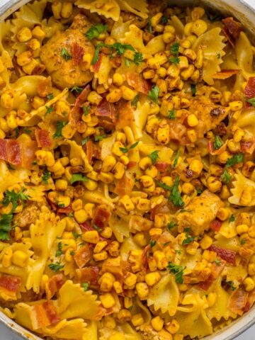 A pot of cooked bowtie pasta mixed with corn, bacon pieces, chicken, and garnished with chopped parsley on a white surface.