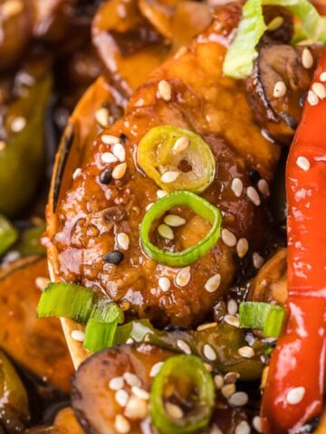 Close-up of a stir-fried dish with chicken, red bell peppers, mushrooms, green onions, and sesame seeds in a savory sauce.