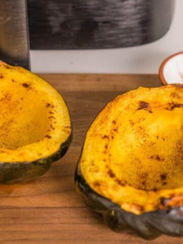 Two halves of cooked acorn squash with cinnamon on a wooden cutting board, next to a fork and a small bowl containing more cinnamon.