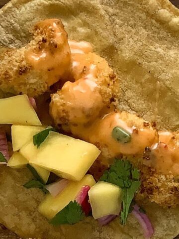 A corn tortilla topped with breaded shrimp, diced mango, red onion, cilantro, and orange sauce on a wooden surface.