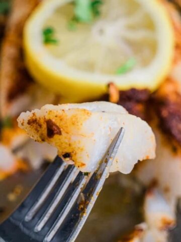 A close-up of a fork holding a bite of seasoned, cooked white fish with sliced lemon and herbs in the background.