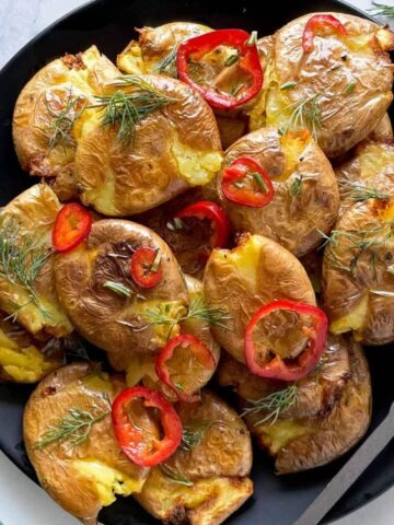 A black plate of smashed roasted potatoes garnished with slices of red chili and fresh dill, with a serving fork beside the plate.