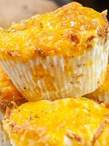 Three cheesy egg muffins in white paper liners are stacked on a plate, with a glass of orange juice and potatoes visible in the background.