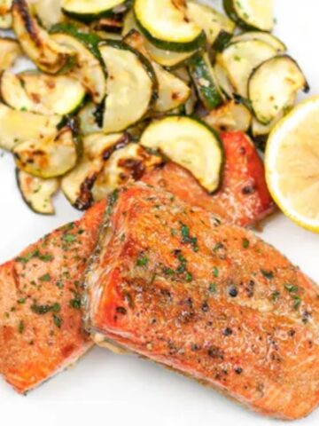 Two pieces of cooked salmon, sautéed zucchini slices, and a halved lemon served on a white plate.