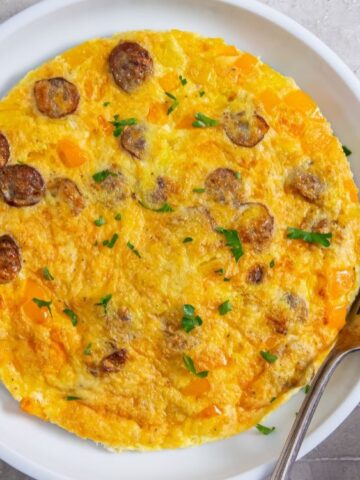 A cooked omelette with sausage slices and diced yellow bell peppers on a white plate, garnished with parsley, with a fork on the side.