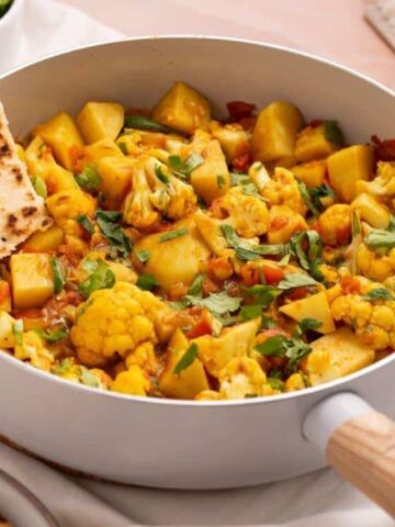 A white pan filled with cooked cauliflower, potatoes, tomatoes, and herbs, garnished with cilantro and served with naan bread on the side.