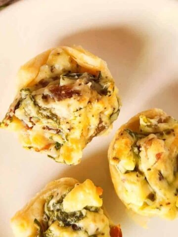 Three mini quiches with herbs and cheese on a white plate, placed on a wooden surface with some greens in the background.
